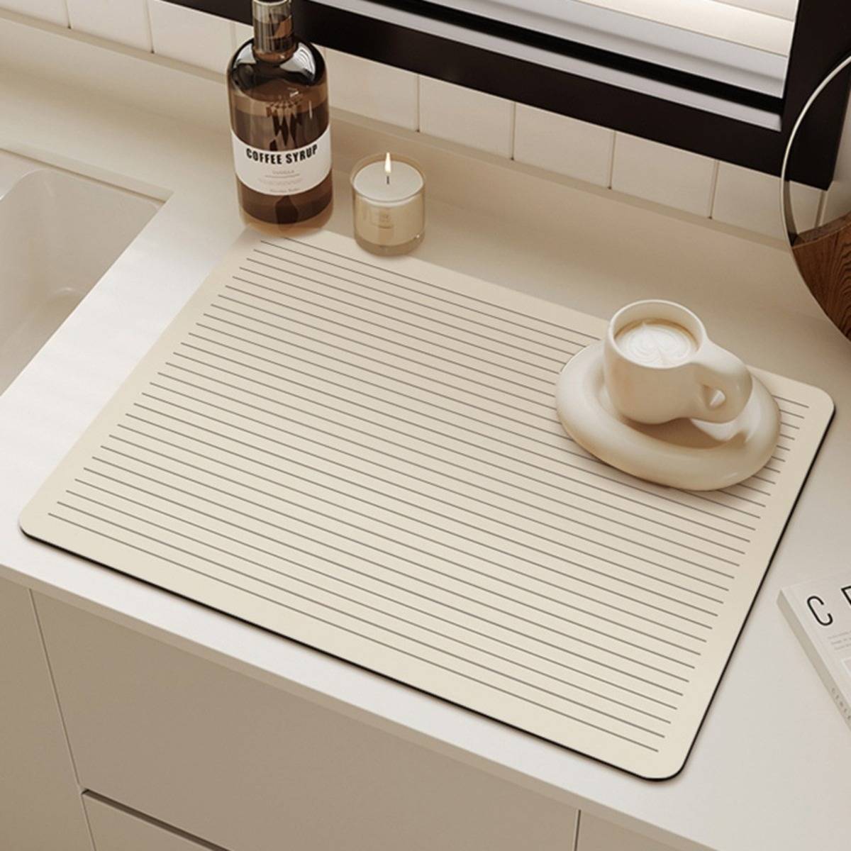 Minimale beige Schreibunterlage auf einer weißen Theke neben einem Fenster, mit einer Kaffeetasse auf einer Untertasse, einer Kaffeesirupflasche und einer kleinen Kerze.