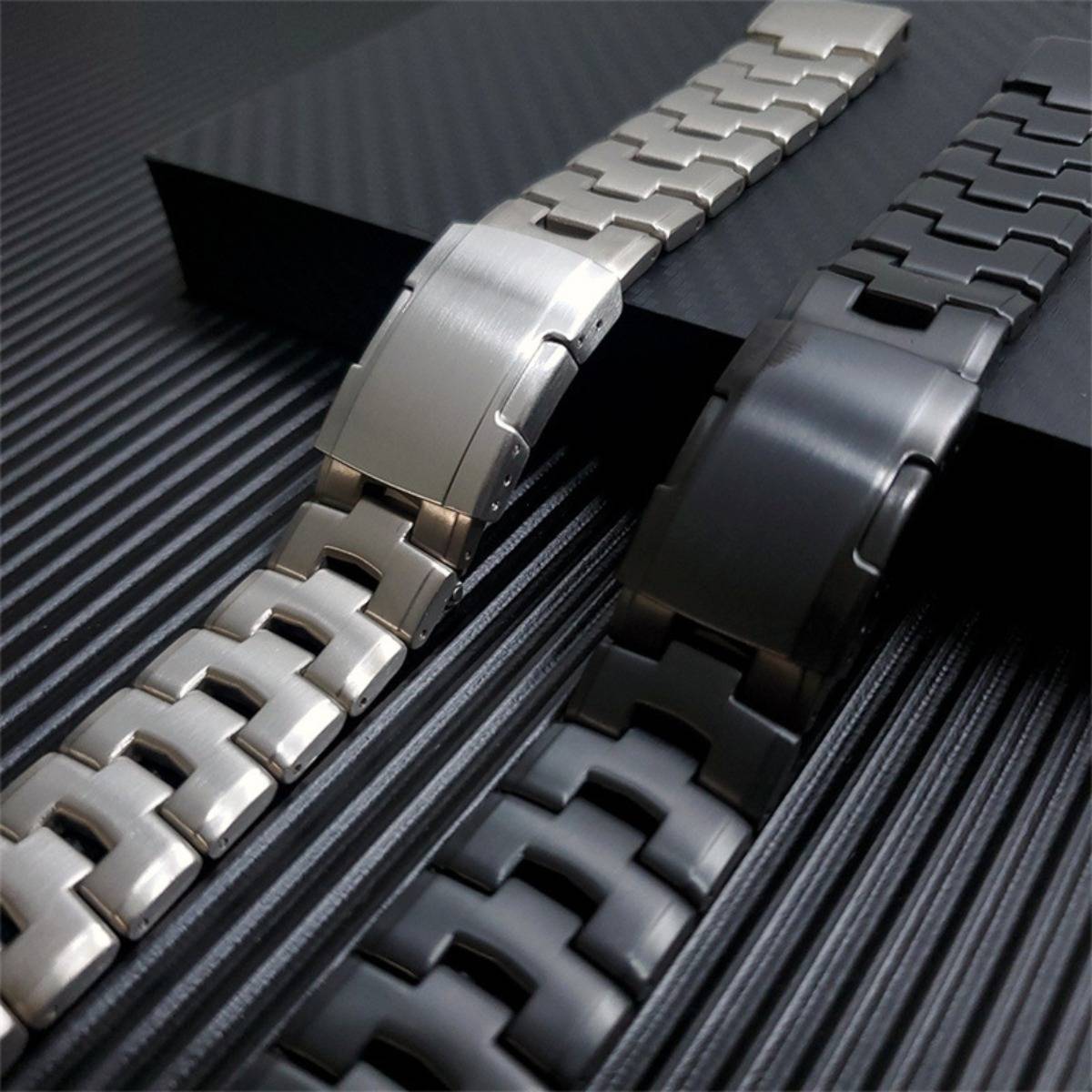 Zwei Metalluhrenarmbänder, eines silberfarben und eines schwarz, nebeneinander auf einer dunkel strukturierten Oberfläche angeordnet.