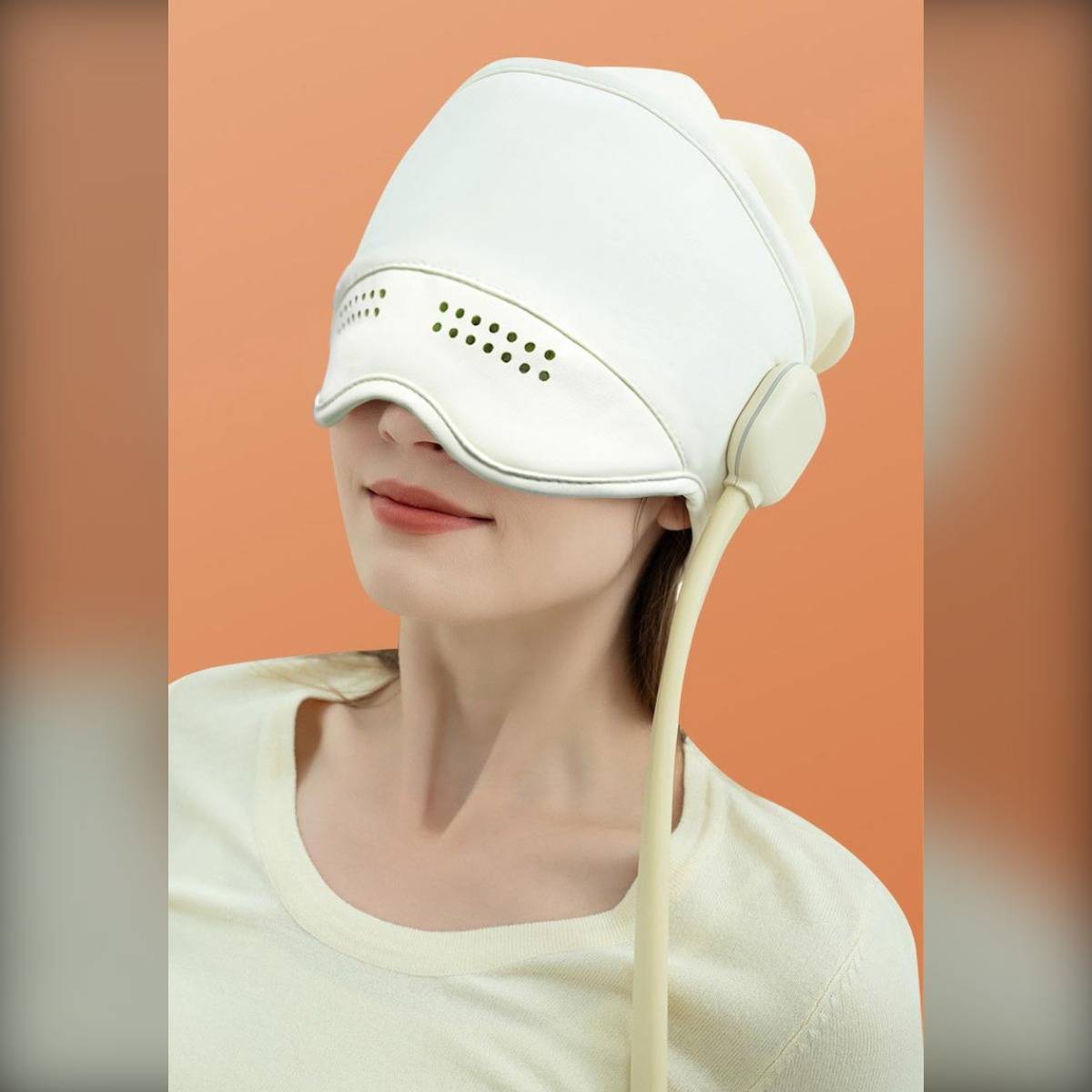 Frau, die ein weißes Augenmassagegerät/eine Gesichtsmaske mit einem Seitenriemen trägt, das ihre Augen vor einem orangefarbenen Hintergrund bedeckt.