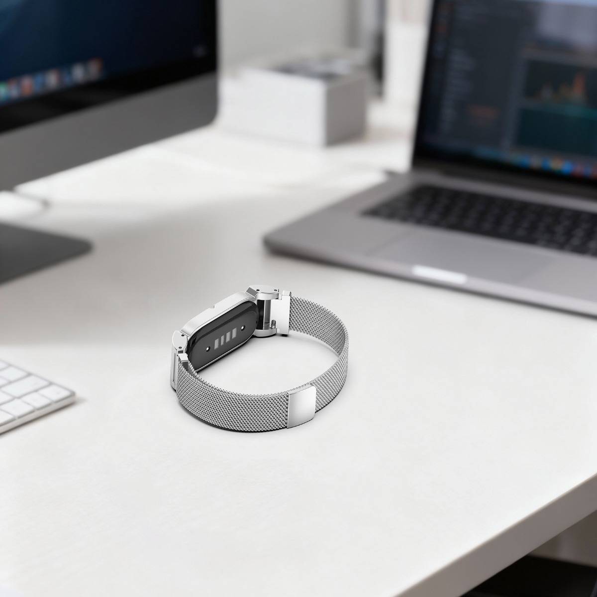 Silberne Smartwatch mit Mesh-Armband auf einem weißen Schreibtisch zwischen zwei aufgeklappten Laptops in einem Büroarbeitsbereich.
