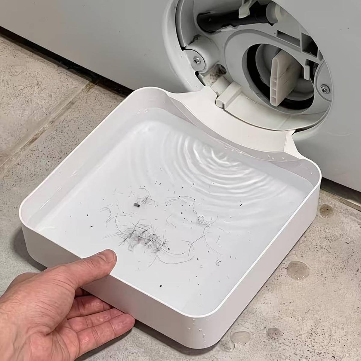 Eine Hand hält ein quadratisches weißes Tablett unter die Ablauföffnung einer Waschmaschine, wobei sich Wasser und Flusen/Schmutz darin gesammelt haben.