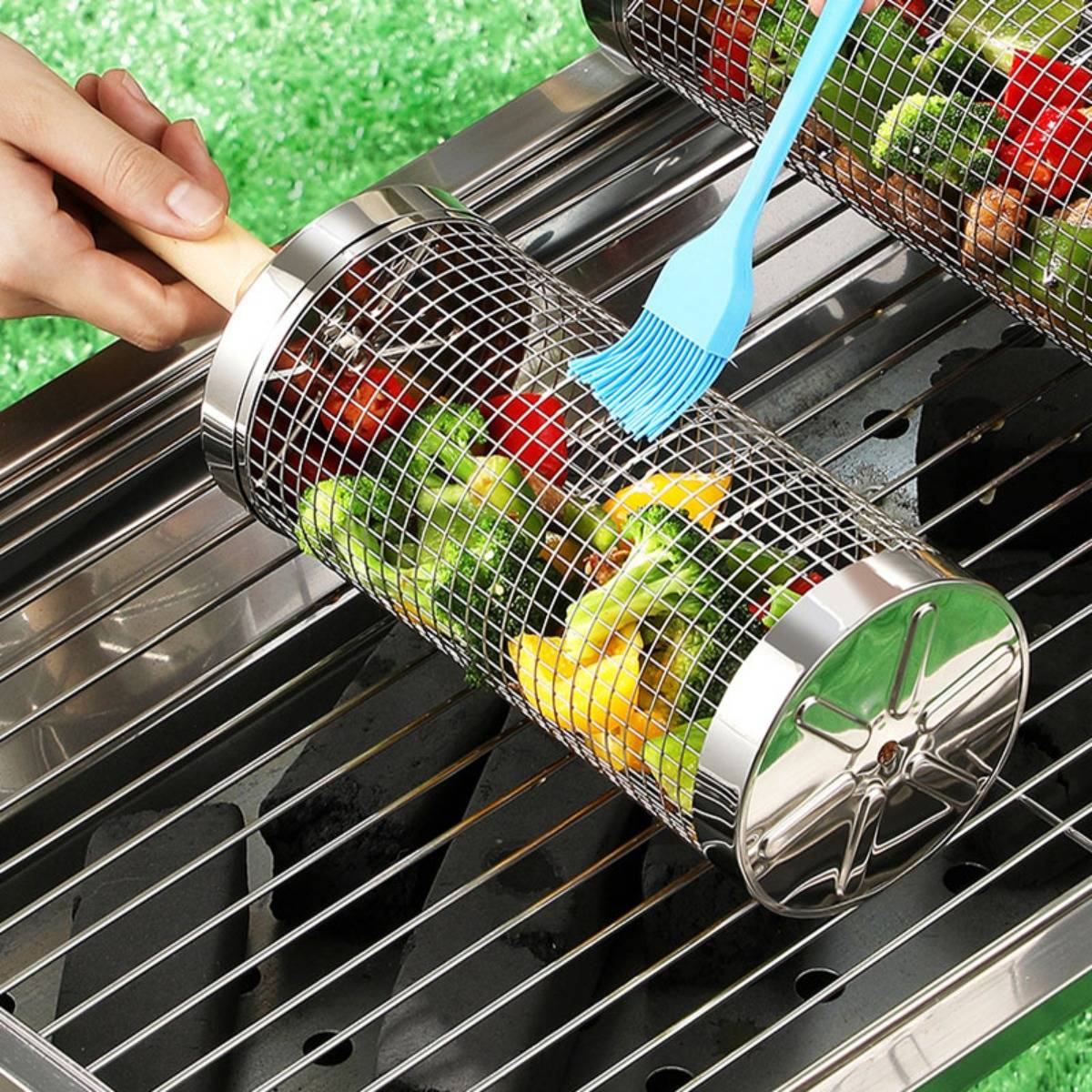 Eine Hand hält einen Metallgrillkorb voller Gemüse über einem Grill, während ein blauer Pinsel Sauce auf das Essen aufträgt.