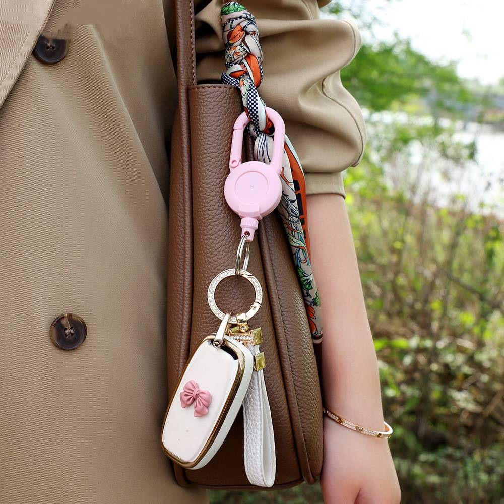 Nahaufnahme eines beigen Mantels und einer braunen Tasche mit einem rosa Schlüsselanhänger mit Ausziehfunktion, der eine weiße Tasche mit Schleifenverzierung und einer weißen Quaste hält.
