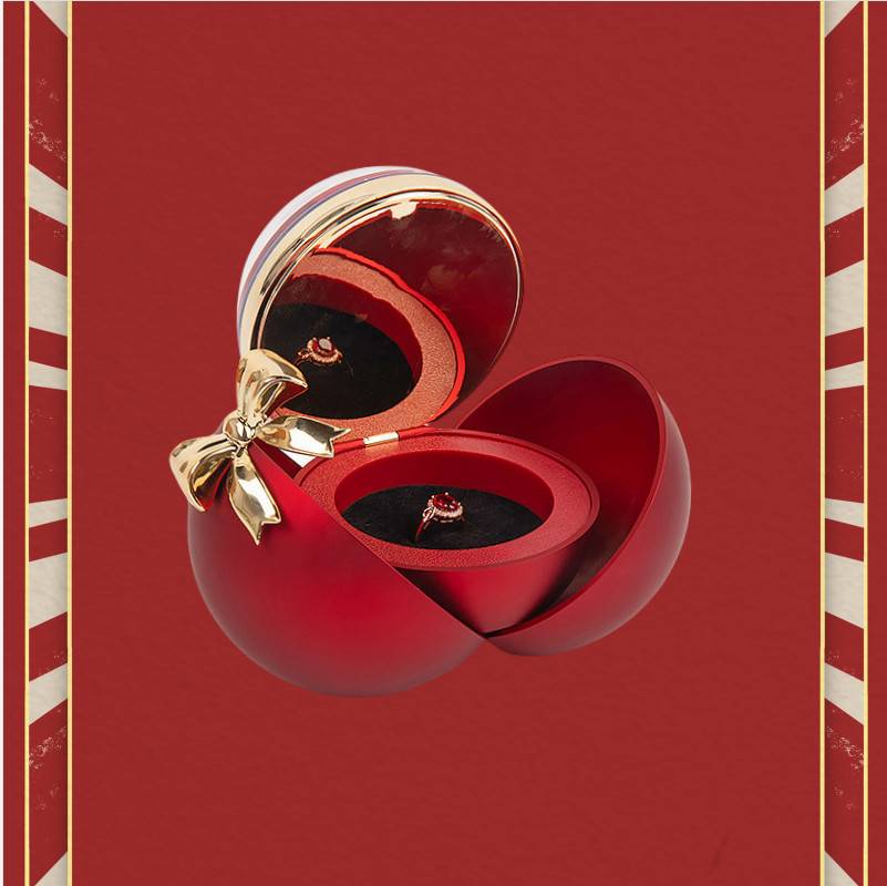 Rote Luxus-Geschenkbox mit einer goldenen Schleife, geöffnet, um eine rote Ringschachtel zu zeigen, die einen Ring vor einem roten Hintergrund mit gestreiften Rändern hält.
