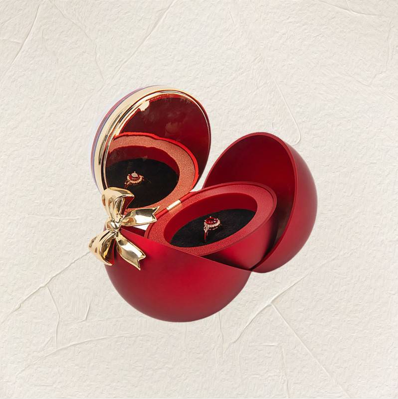 Offene herzförmige rote Schmuckschatulle mit goldener Schleife, enthaltend einen Ring mit einem roten Edelstein auf einem hellstrukturierten Hintergrund.