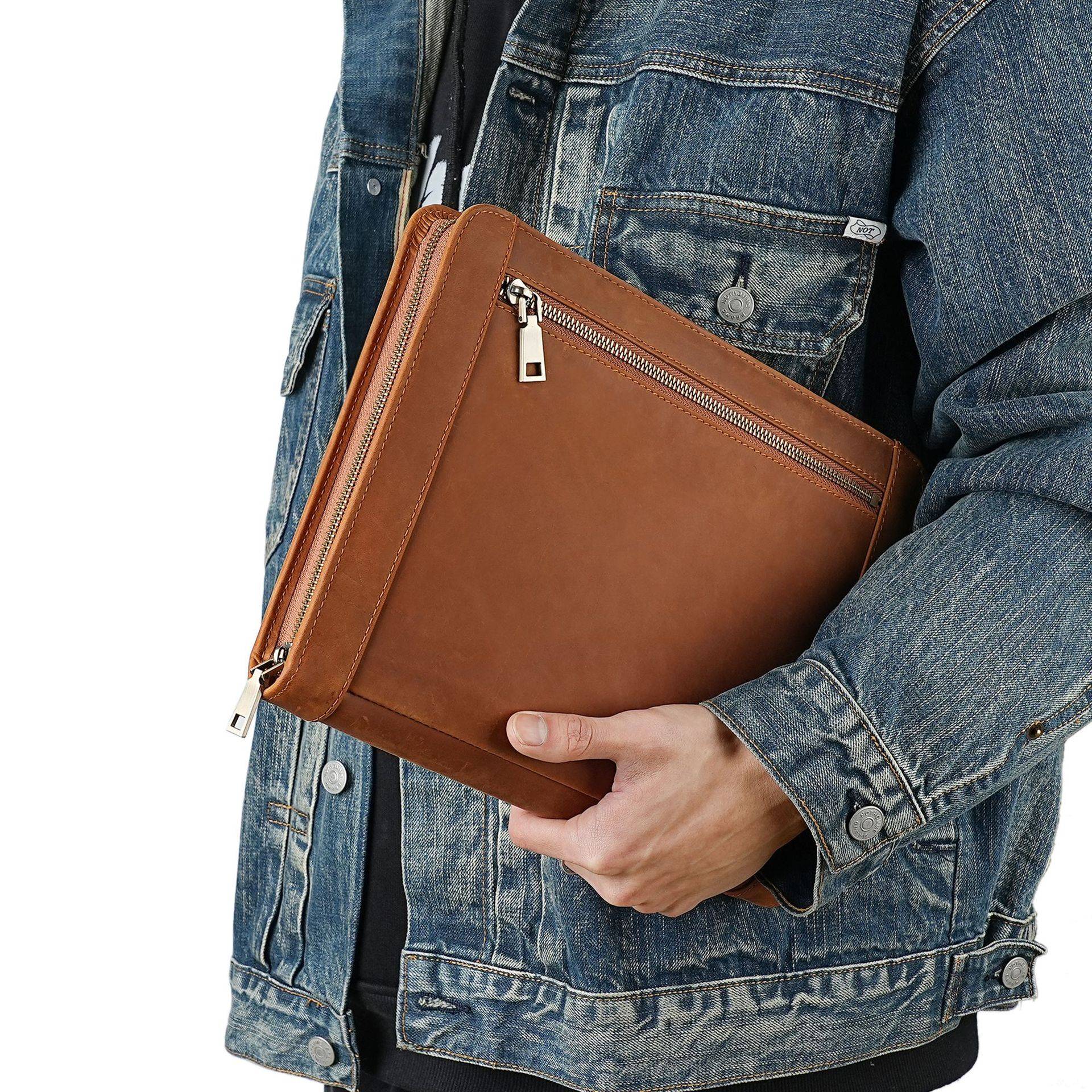 Person in a Jeansjacke hält ein braunes Lederportfolio mit Reißverschluss oder eine Tablet-Tasche gegen einen weißen Hintergrund.