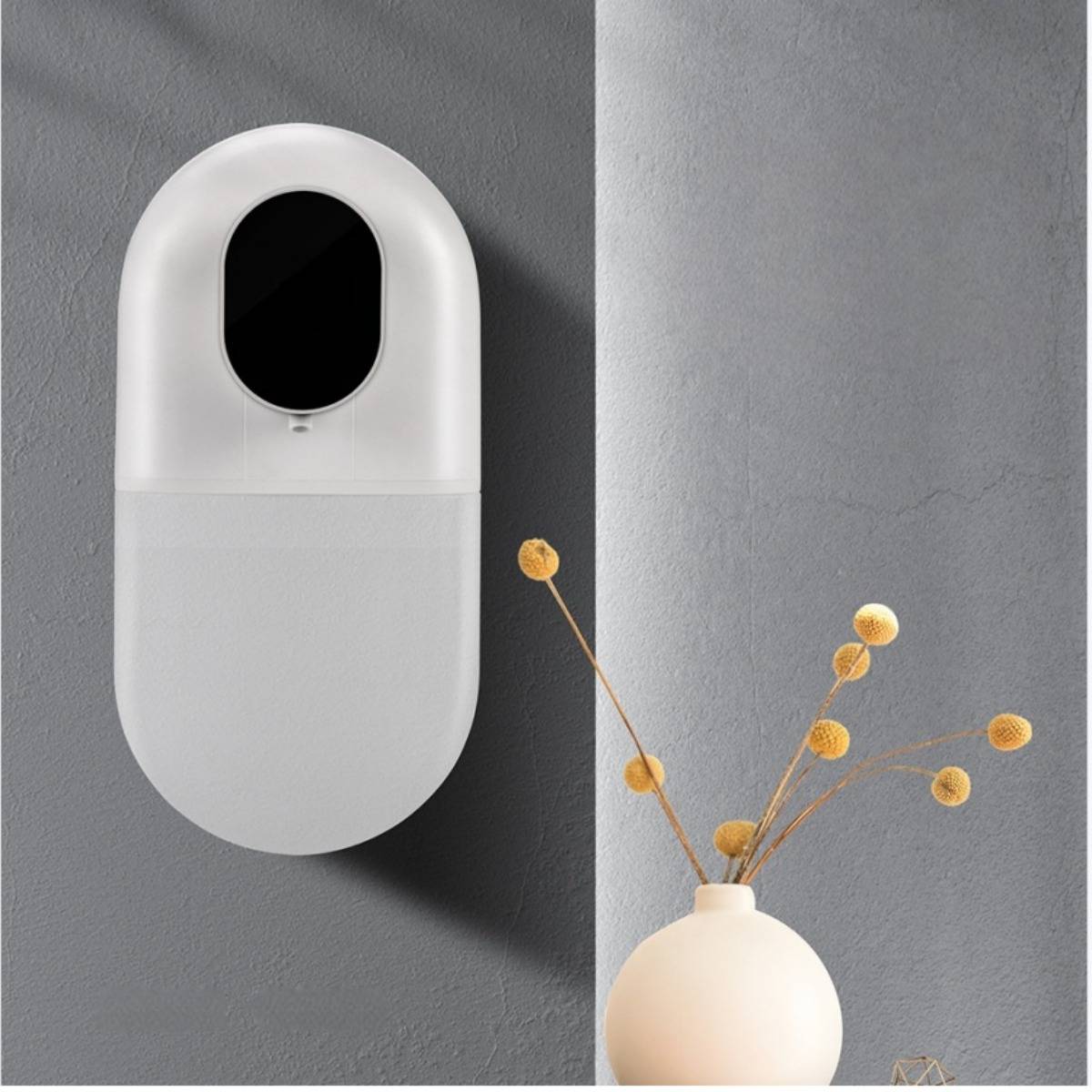 Wandmontierter ovaler weißer Spender oder Lufterfrischer an grauer Wand neben einer kleinen Vase mit getrockneten Stielen.