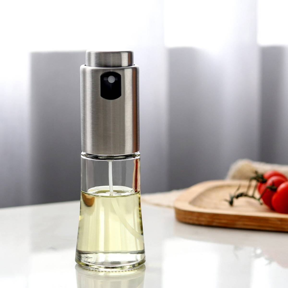 Edelstahl- und Glasöldispenserflasche auf einem Tisch, mit unscharfen Tomaten im Hintergrund.