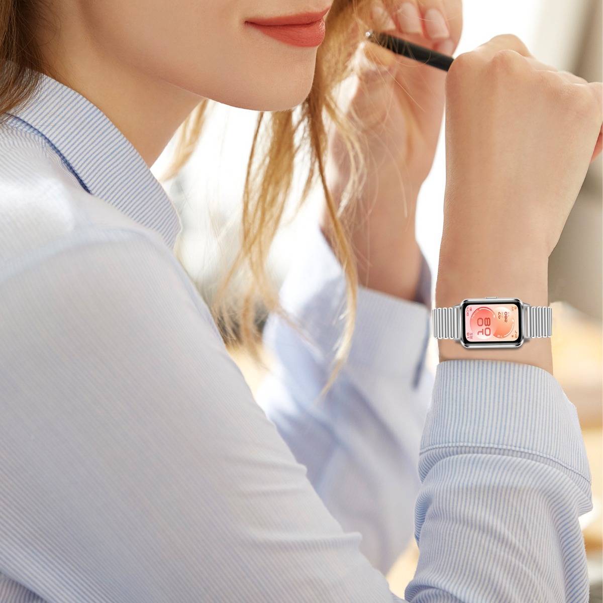 Nahaufnahme einer Frau, die ihr Haar zurechtmacht und eine silberne Smartwatch mit einem rosa digitalen Zifferblatt an ihrem Handgelenk trägt.