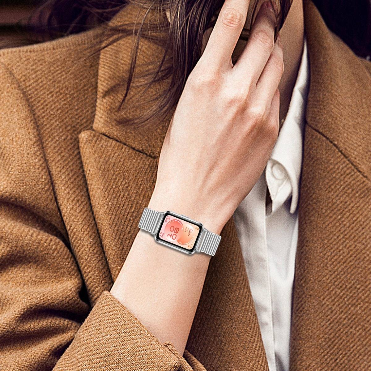 Nahaufnahme einer Person, die eine rechteckige silberne Smartwatch mit rosa digitalem Display trägt, kombiniert mit einem braunen Mantel und weißem Hemd.