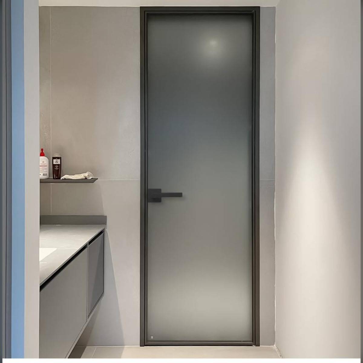 Minimalistische Badezimmereinrichtung mit Glastür aus Mattglas, Waschtischplatte und kleinem Wandregal mit Toilettenartikeln.