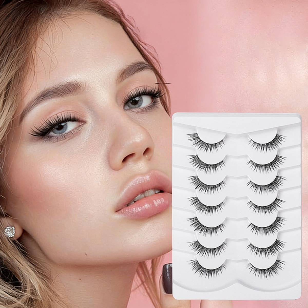 Nahaufnahme einer Frau mit langen Wimpern neben einem Tablett mit Paaren von künstlichen Wimpern auf rosa Hintergrund.