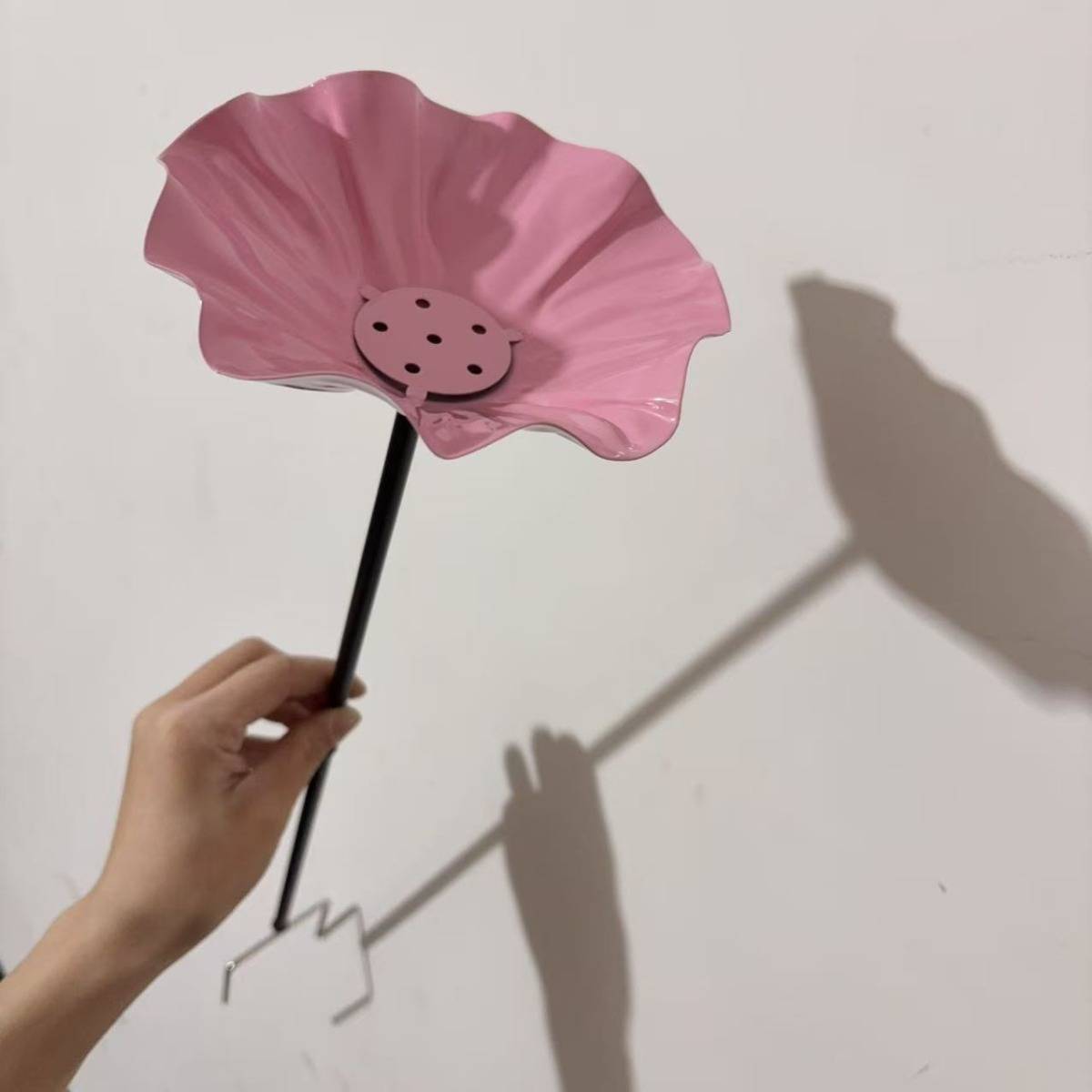 Hand hält ein rosa blütenförmiges Papierhandwerk auf einem Stab, das einen Blumenschatten auf eine Wand wirft.