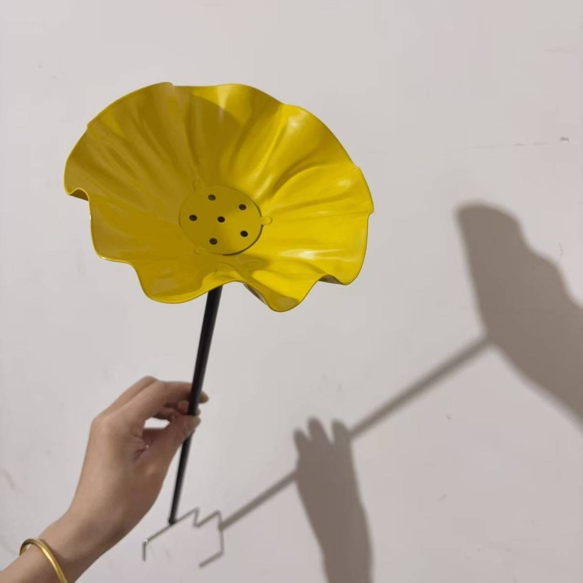 Eine Hand hält einen gelben blumenförmigen Metallgartenstab oder Windspinner mit schwarzem Stiel, der einen Schatten auf einer Wand wirft.