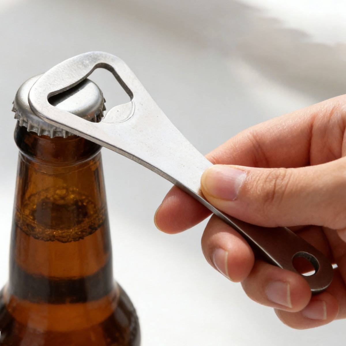 Hand benutzt einen Metall-Flaschenöffner, um den Deckel einer bernsteinfarbenen Glassbierflasche abzuhebeln.