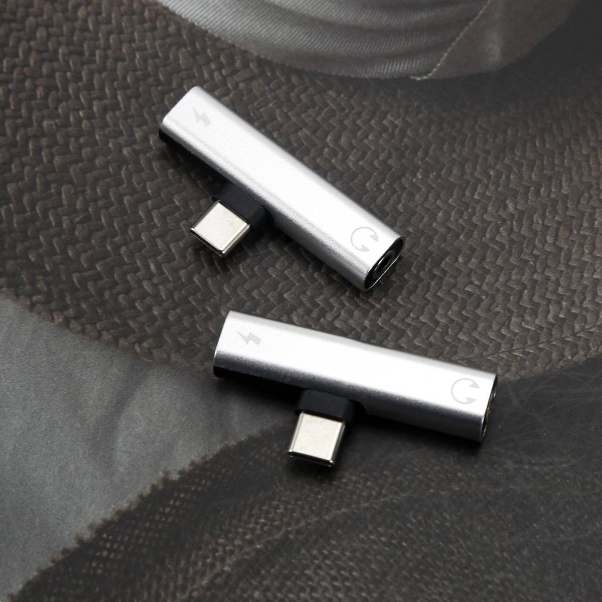 Zwei silberne USB-C-Adapter-Dongles auf einer dunklen strukturierten Oberfläche, jeweils mit einem kleinen USB-C-Stecker und einem abgerundeten Ende, nebeneinander dargestellt.