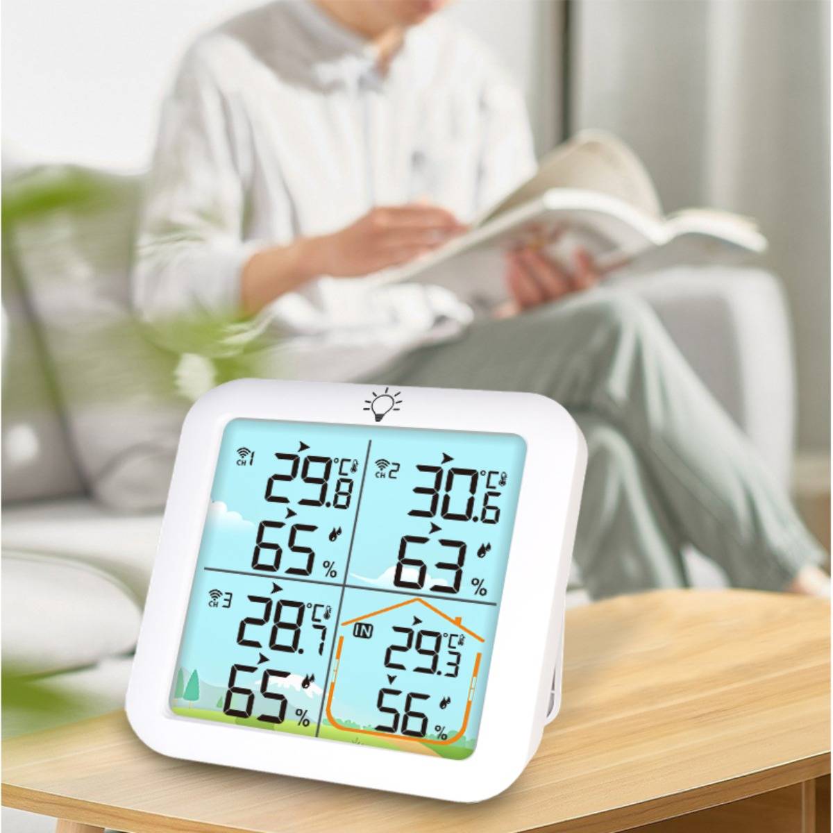 Digitales Innenthermometer-Hygrometer auf einem Tisch zeigt Raumtemperatur- und Feuchtigkeitswerte an, mit einer unscharfen Person, die im Hintergrund liest.