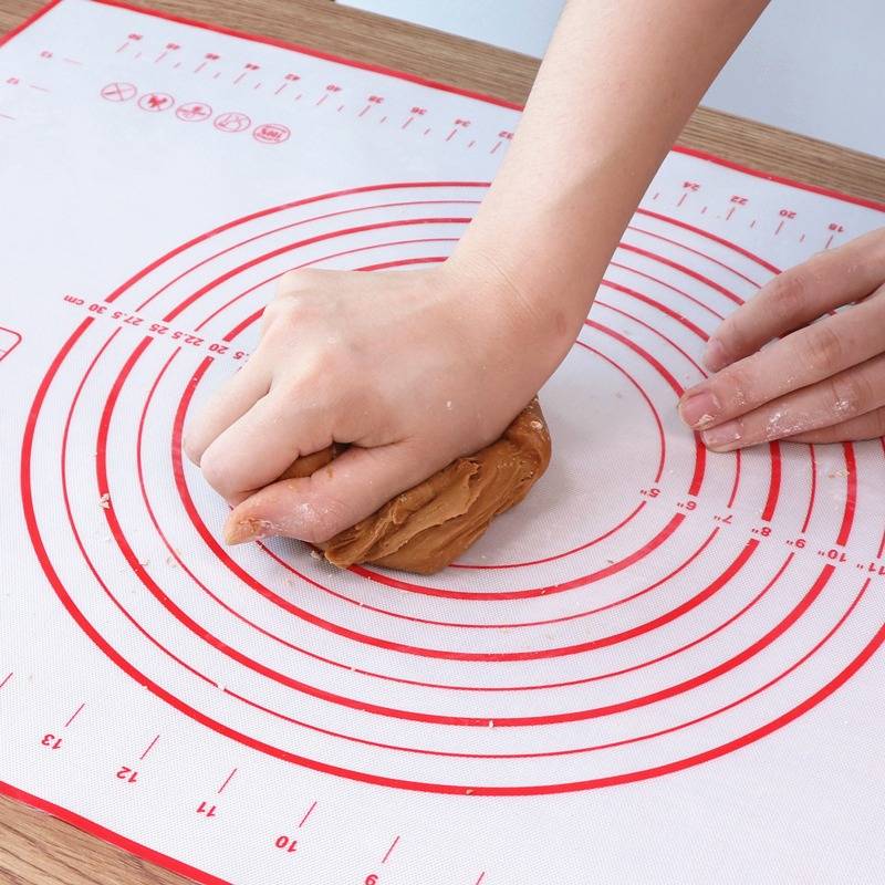 Hände kneten braunen Teig auf einer rot umrandeten Silikonbackmatte auf einem Holztisch.