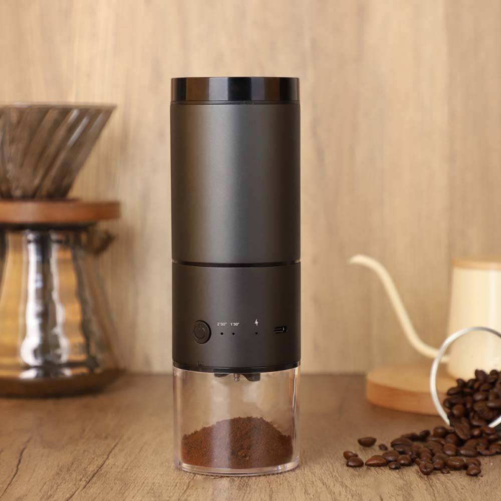 Elektrische Kaffeemühle mit Kaffeemehl in einem durchsichtigen Behälter, dargestellt auf einer Holztheke mit Kaffeebohnen in der Nähe.