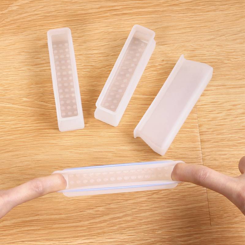 Hände dehnen eine flexible, durchscheinende Silikonhülle über ein rechteckiges Objekt, während drei ähnliche Hüllen auf einem Holztisch liegen.