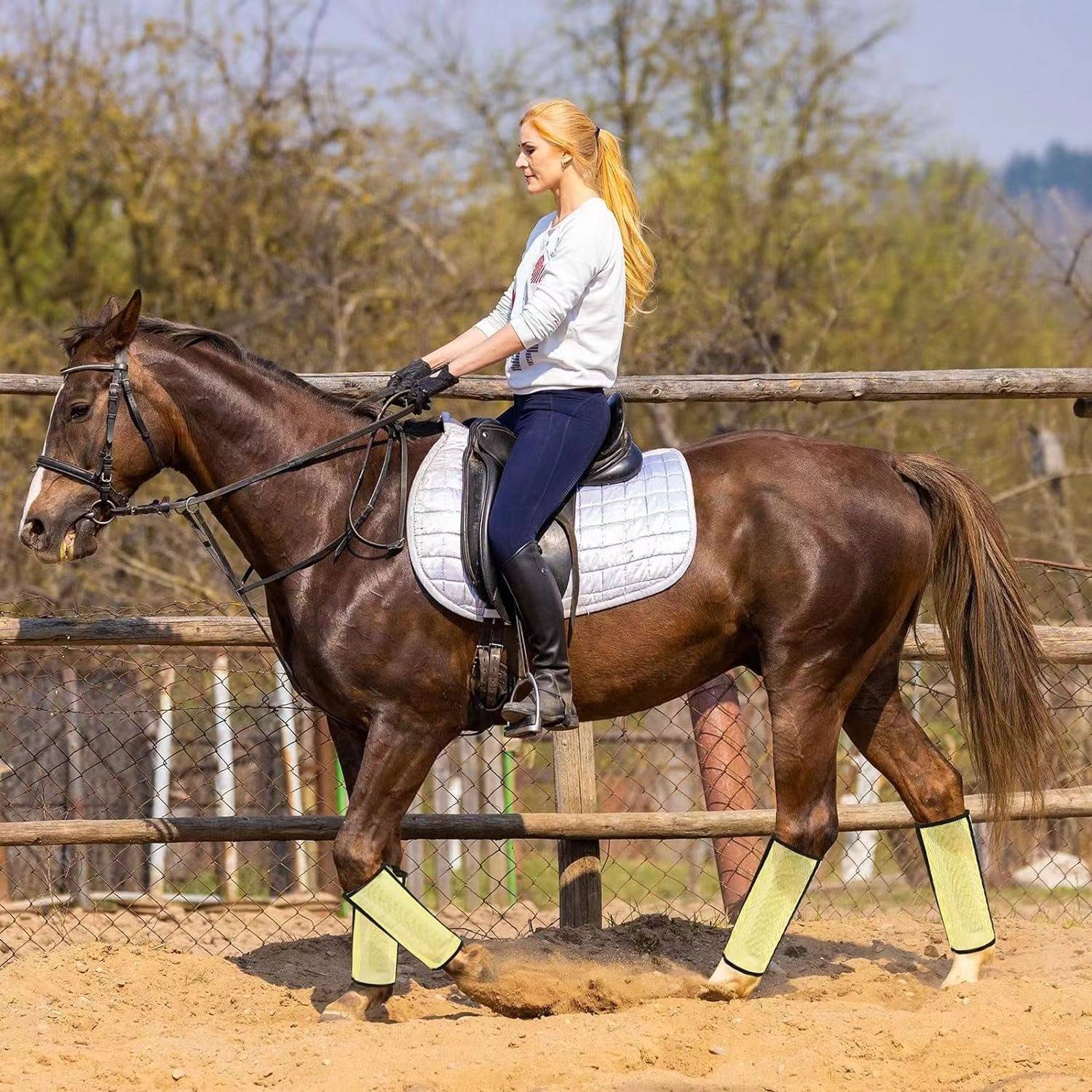 Frau auf einem braunen Pferd in einer sandigen Außenreitanlage reitend, gekleidet in Reitkleidung und gelben Gamaschen.