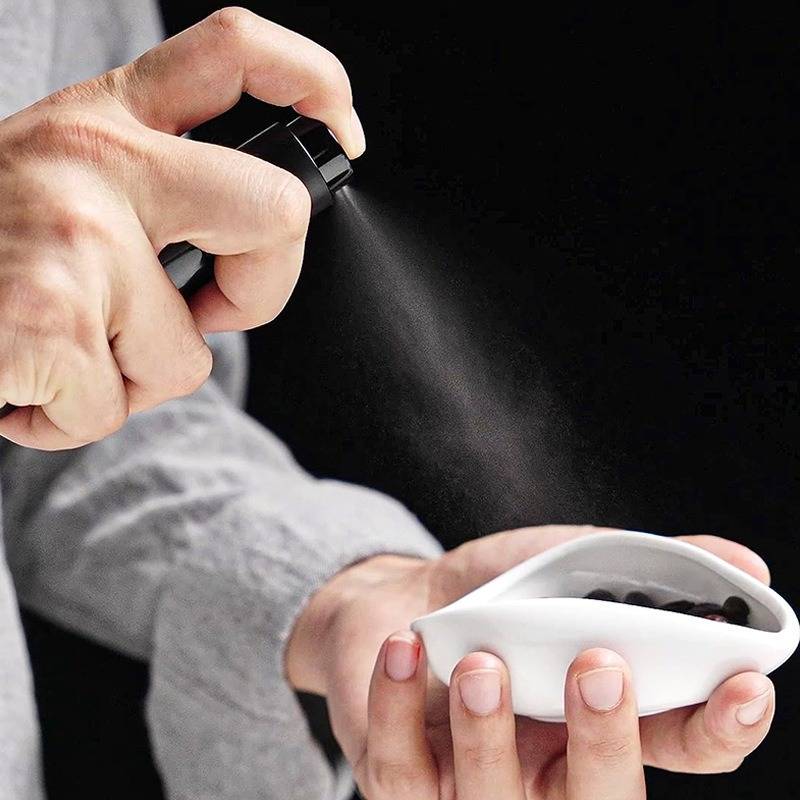 Eine Person sprüht Flüssigkeit aus einer schwarzen Flasche in eine kleine weiße Schale, die sie in ihrer anderen Hand hält.