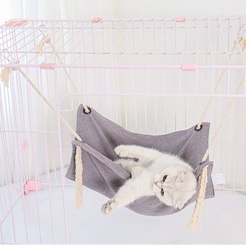 Weiße Katze, die in einer grauen Hängematte in einem weißen Käfig liegt, seitlich ruhend mit entspannten Pfoten.