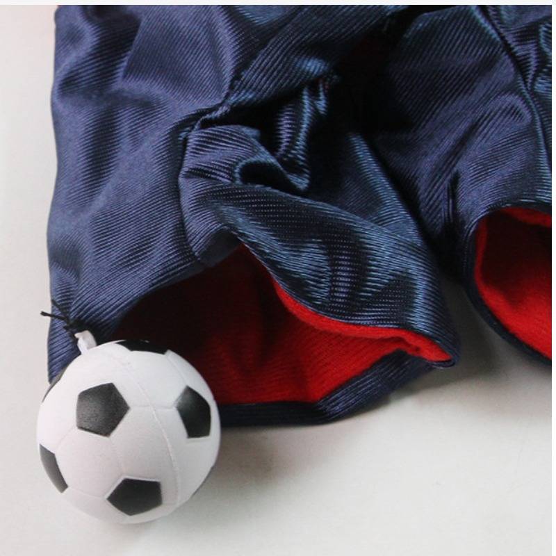 Ein kleiner Fußball, der teilweise unter einem dunkelblauem Stoff mit roter Fütterung auf einer weißen Oberfläche verstaut ist.