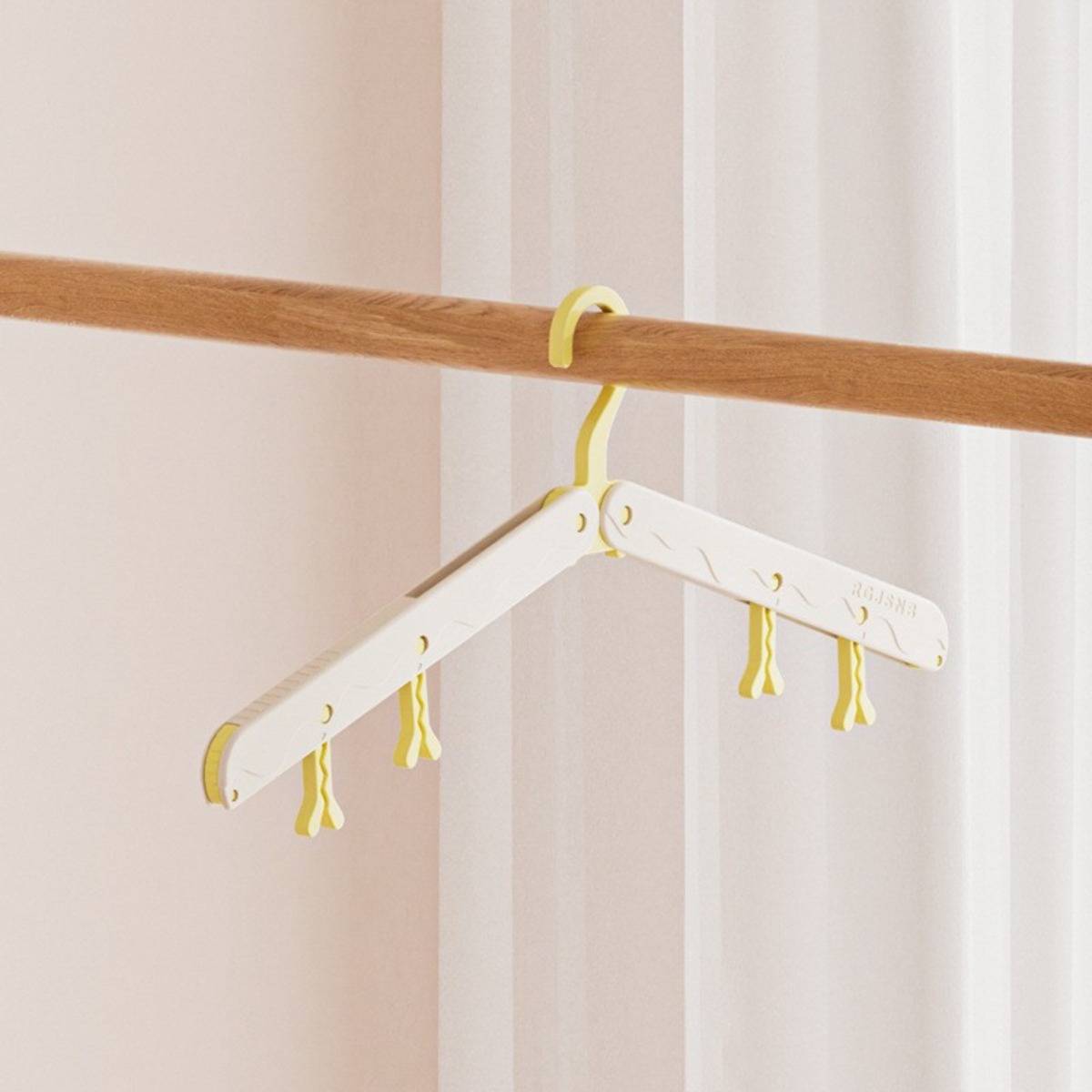 Weiße klappbare Kleiderbügel mit gelben Akzenten, die auf einer Holzstange vor transparenten Vorhängen hängen.