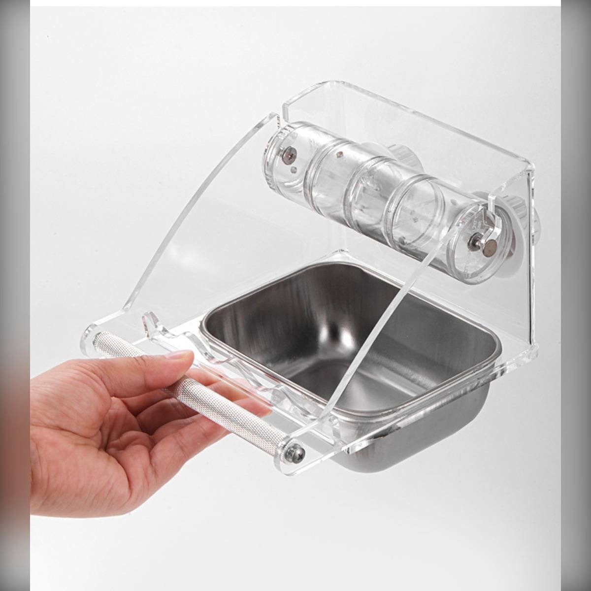 Hand hält einen durchsichtigen Kunststoff-Haustierfutterautomat mit einer Edelstahlschüssel und angebrachtem Wasserspender, gezeigt vor weißem Hintergrund.