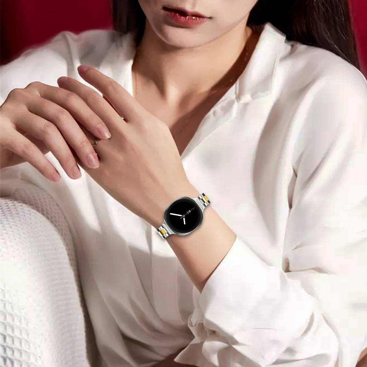 Frau, die eine schwarze runde Armbanduhr mit einem silber-goldenen Armband trägt, gezeigt auf ihren verschränkten Händen vor einem roten Hintergrund.