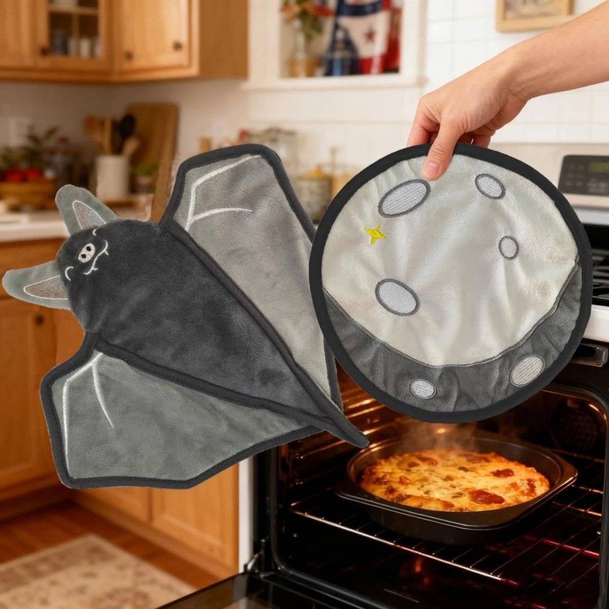 Eine Hand hält neuartige Ofenhandschuhe in Form einer Fledermaus und eines Mondes vor einem offenen Ofen mit einer Pizza, die darin backt.
