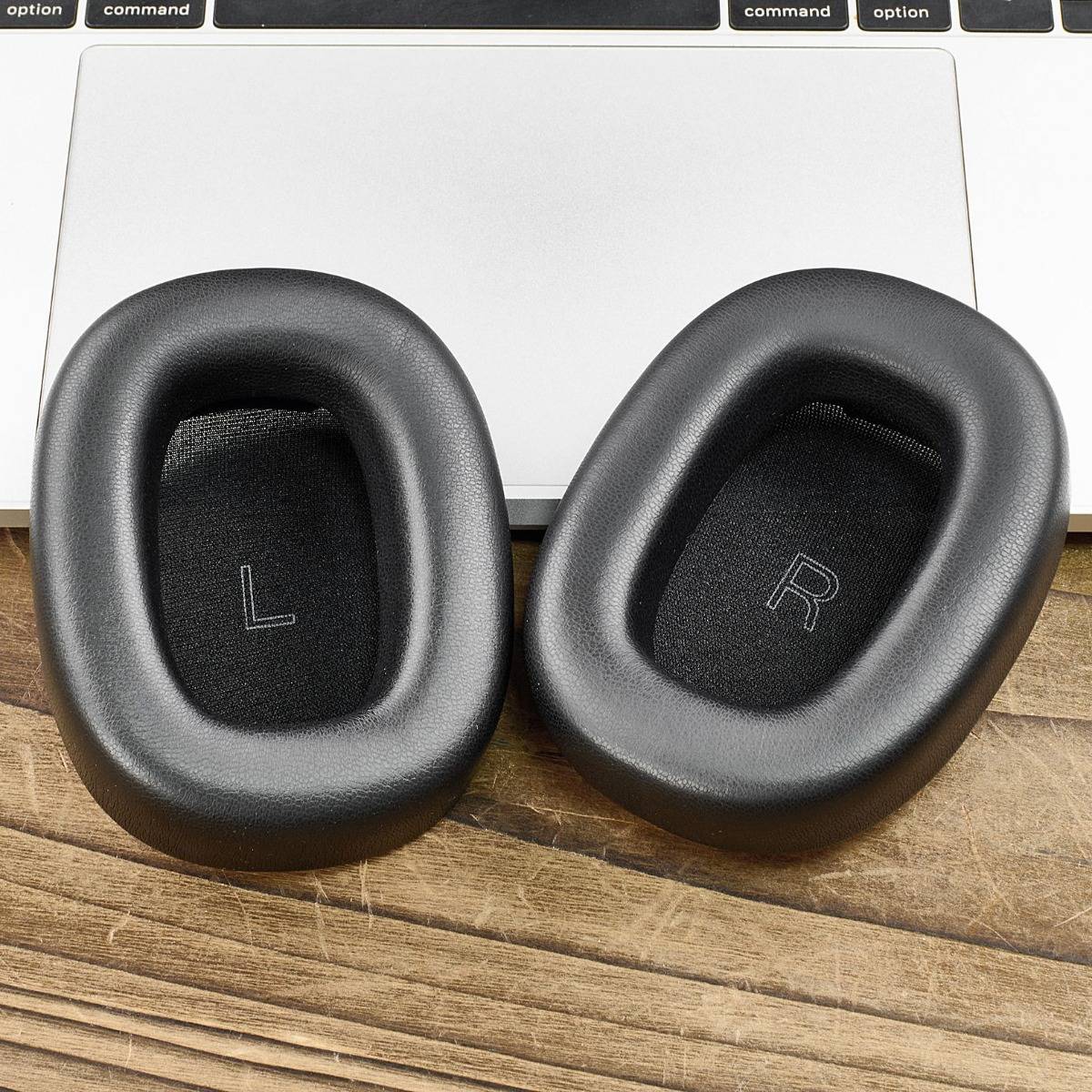 Paar schwarze Over-Ear-Kopfhörer-Ohrpolster mit den Bezeichnungen L und R, gezeigt neben einer Laptop-Tastatur auf einer Holzoberfläche.