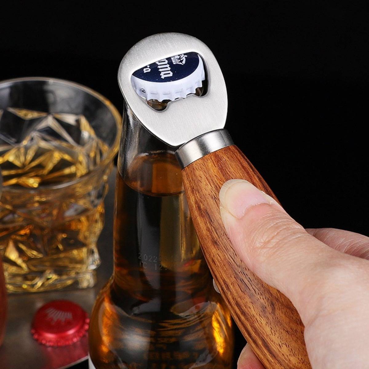 Hand benutzt einen Flaschenöffner mit Holzgriff, um einen Bierflaschenverschluss neben einem Glas Whiskey auf dunklem Hintergrund abzuhebeln.