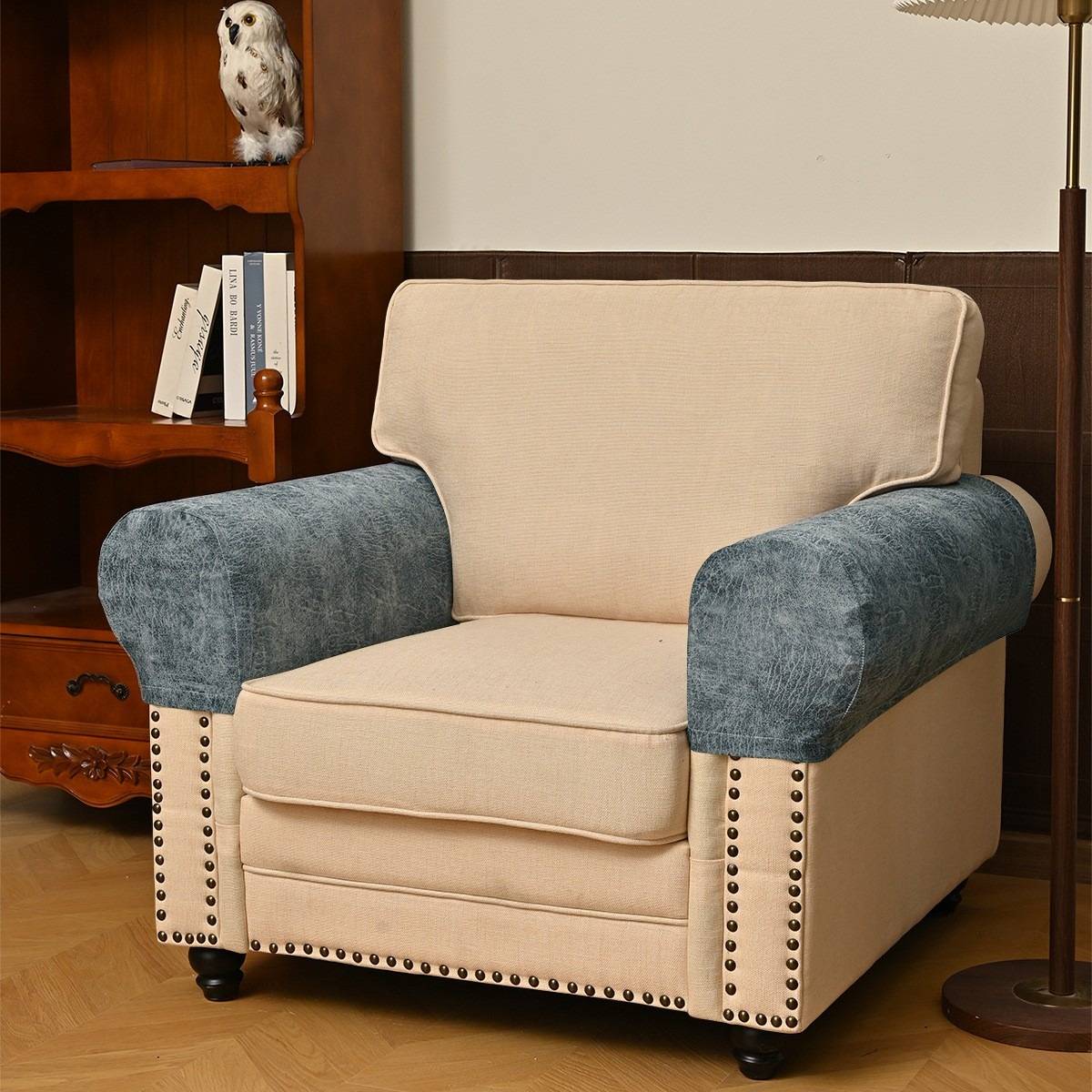 Beige gepolsterter Sessel mit blauen Armlehnen und Nagelkopfverzierung in einem Wohnzimmer, neben einem Holzbücherregal und einer Stehlampe.