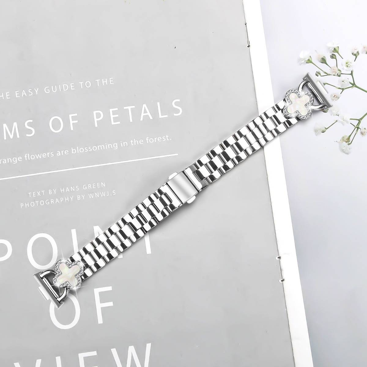 Silber-und-schwarz gestreifter Armbanduhr mit blumenförmigen Akzenten, diagonal auf einem grauen Buchcover neben weißen Blüten angezeigt.