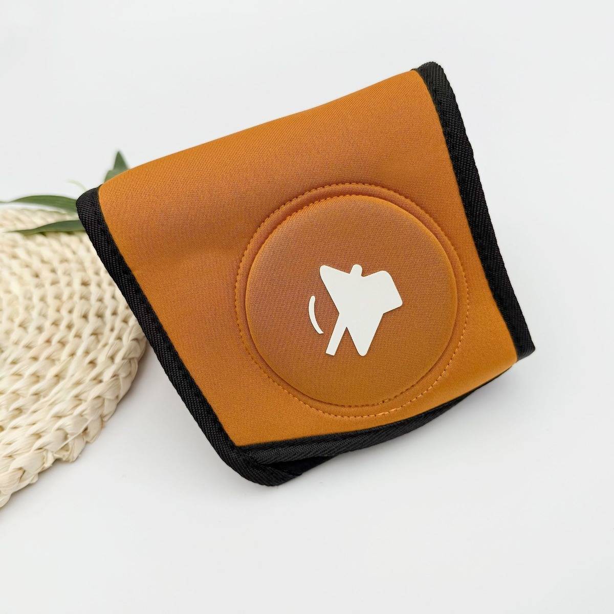 Orange-schwarze gefaltete Geldbörse oder Tasche mit weißem Pferdelogo, gezeigt auf einer weißen Oberfläche neben einem geflochtenen Korb.