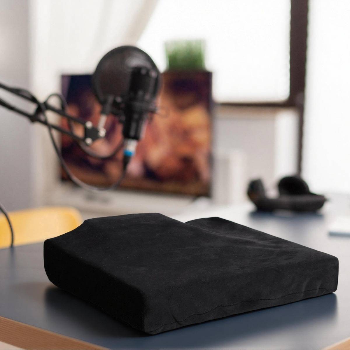 Schwarzes ergonomisches Sitzkissen auf einem Schreibtisch mit einem Mikrofon im Hintergrund, was auf ein Heim-Podcast- oder Aufnahme-Setup hindeutet.