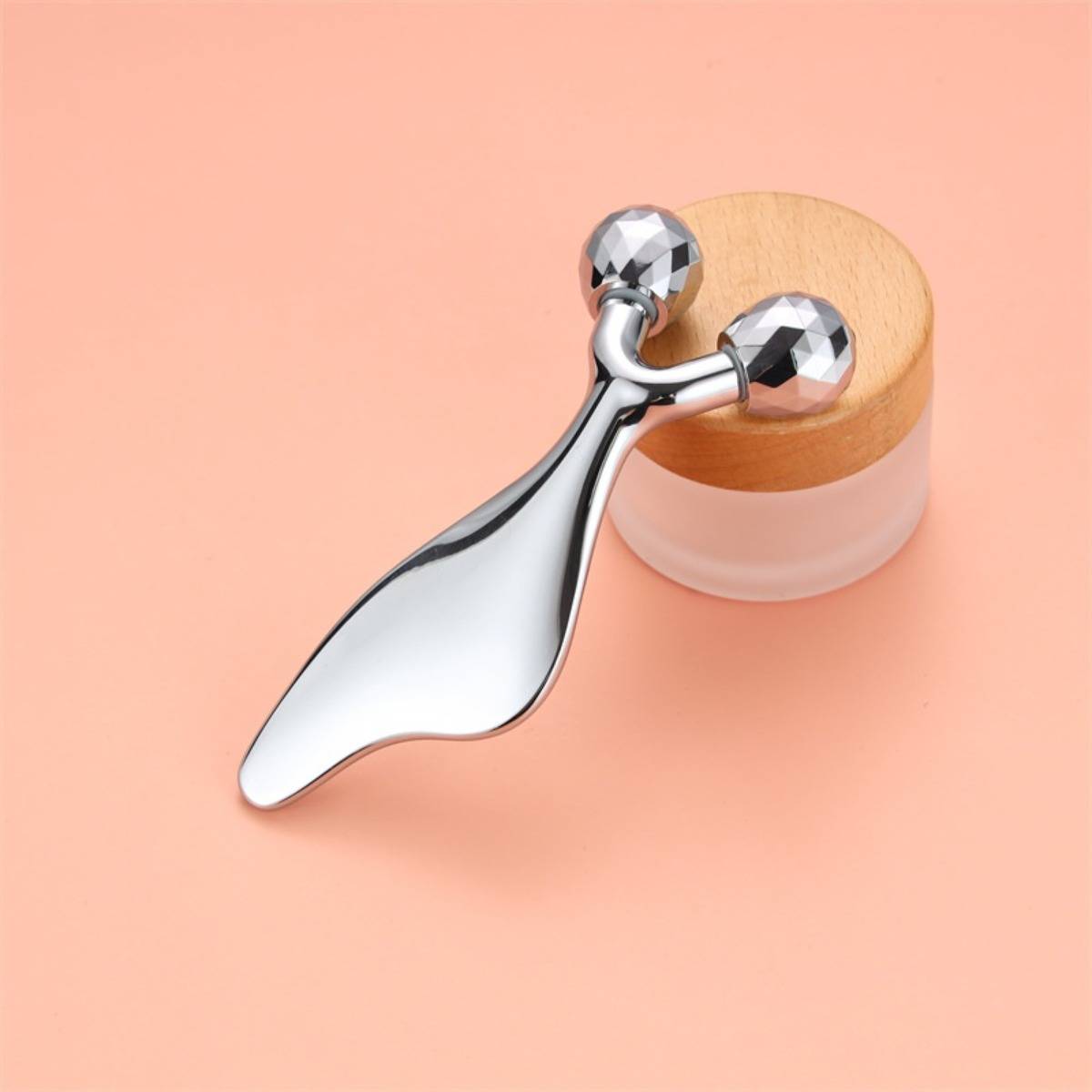 Chrome-Gesichtsmassageroller mit zwei facettierten Kugeln, die auf einem kleinen runden Glas auf pfirsichfarbenem Hintergrund ruhen.