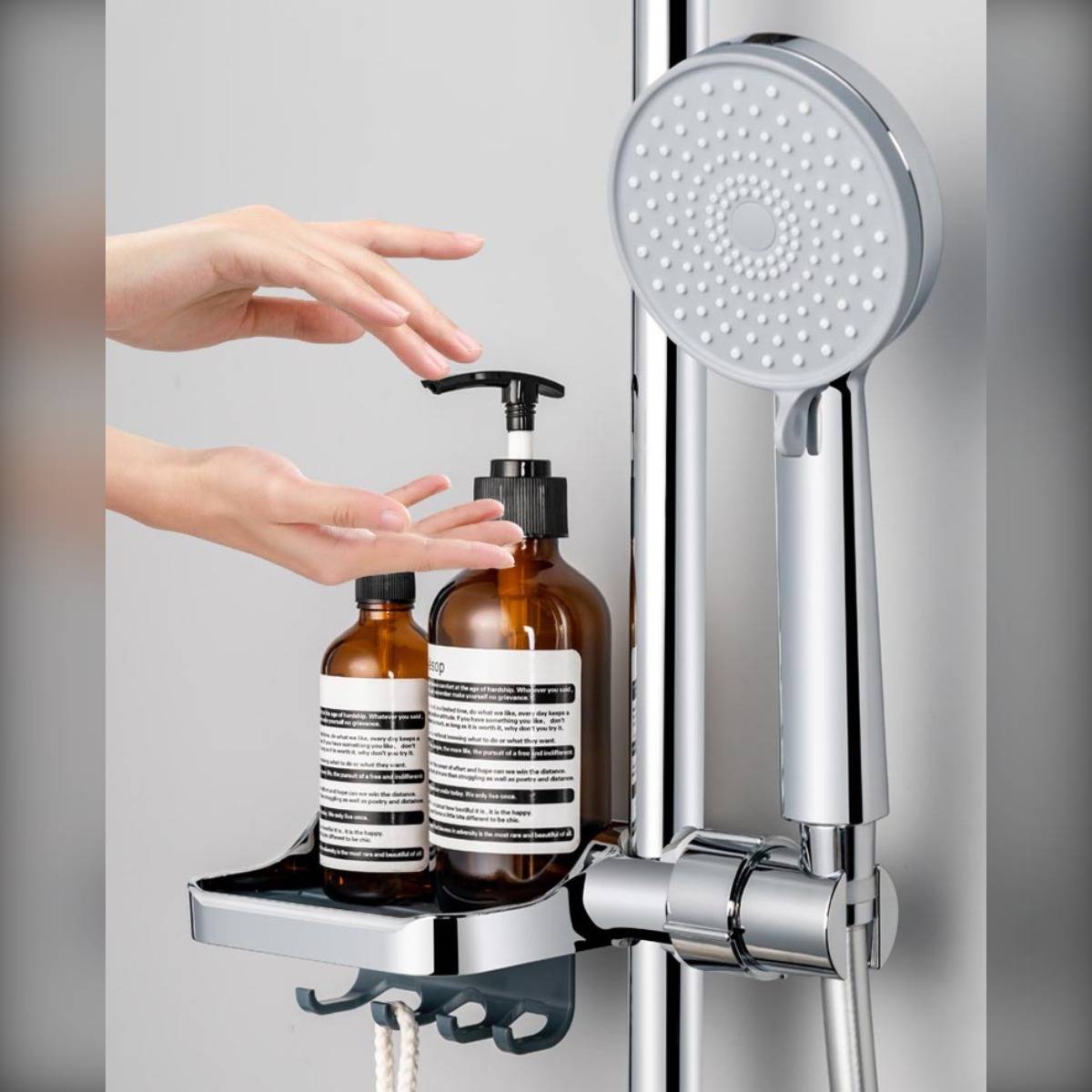 Hände, die eine Pumpflasche und eine kleinere Flasche auf einem verchromten Duschablage neben einem Handbrausekopf in einem Badezimmer platzieren.