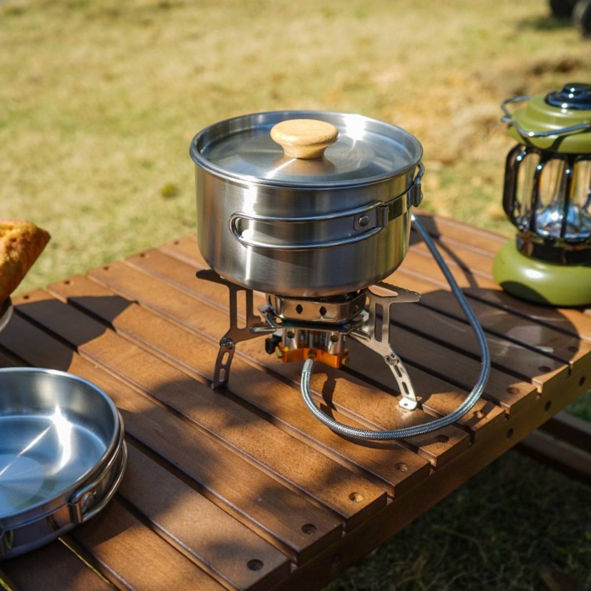 Tragbarer Campingkocher mit einem Topf mit Deckel aus Metall auf einem Holztisch, neben einer Laterne und Kochutensilien im Freien.
