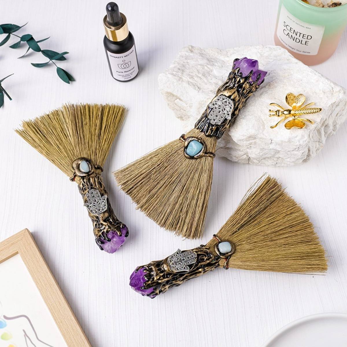 Drei dekorative Handfeger mit violetten Kristallgriffen und Boho-Anhängern auf einer weißen Oberfläche angeordnet, zusammen mit Kerzen, Blättern und kleinen Dekorelementen.