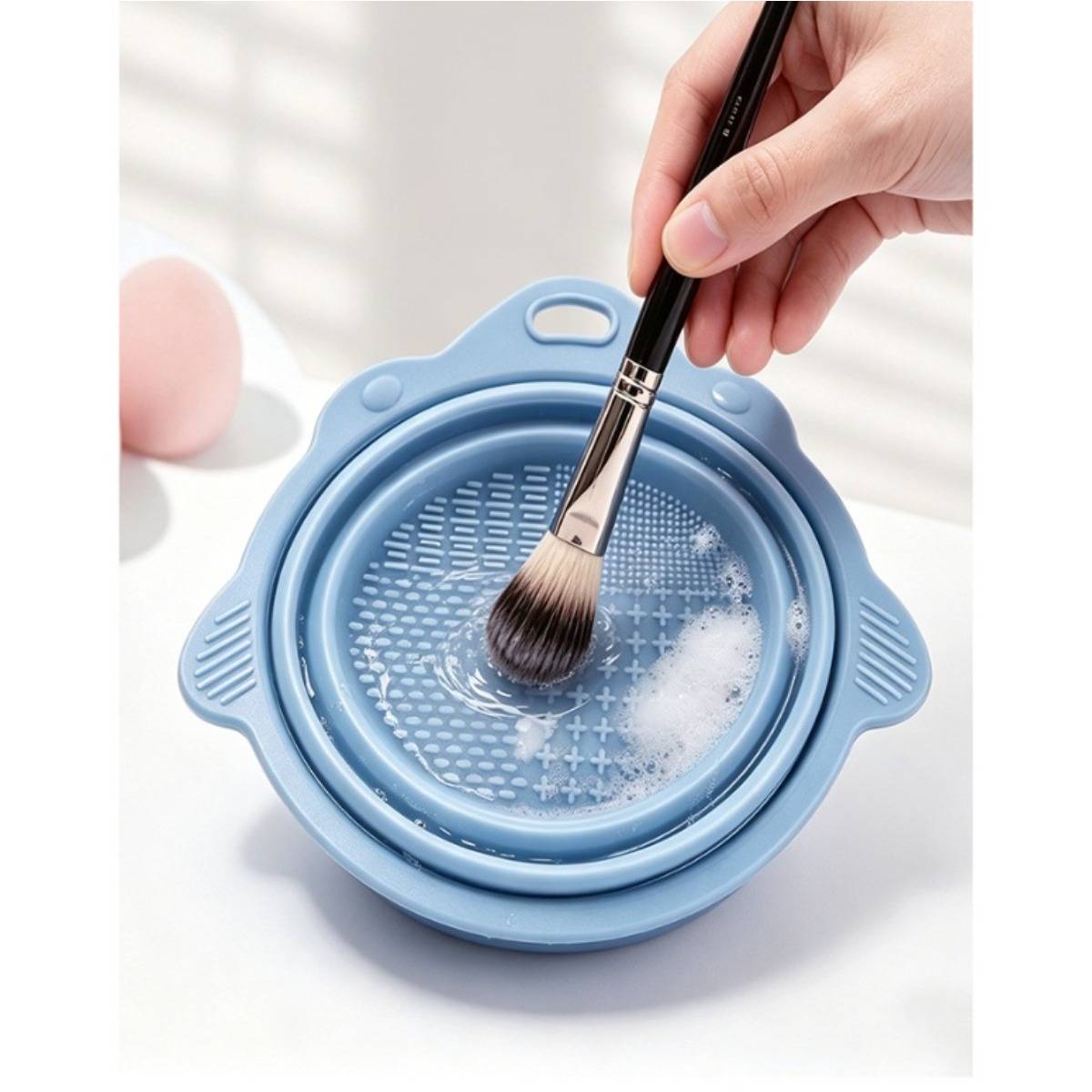 Eine Make-up-Bürste mit der Hand in einer blauen Silikonbürsten-Reinigungsschale mit Seifenschaum reinigen.
