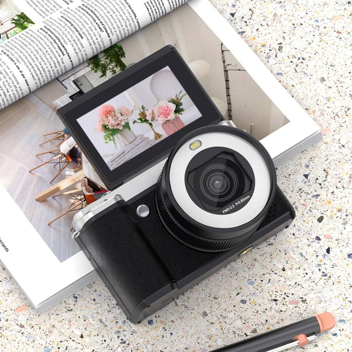 Digitalkamera mit hochklappbarem Bildschirm, auf dem eine Blumengestaltung zu sehen ist, die auf einer aufgeschlagenen Zeitschrift neben einem Bleistift ruht.