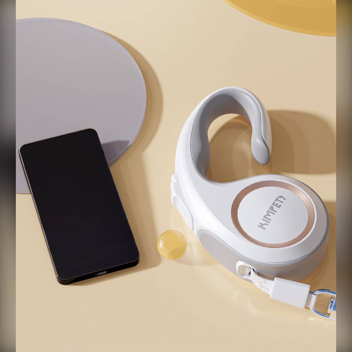 Smartphone neben einem weißen Over-Ear-Kopfhörer auf beigem Hintergrund, mit einer runden Schutzhülle oder einem Kissen und kleinen gelben Akzentobjekten.