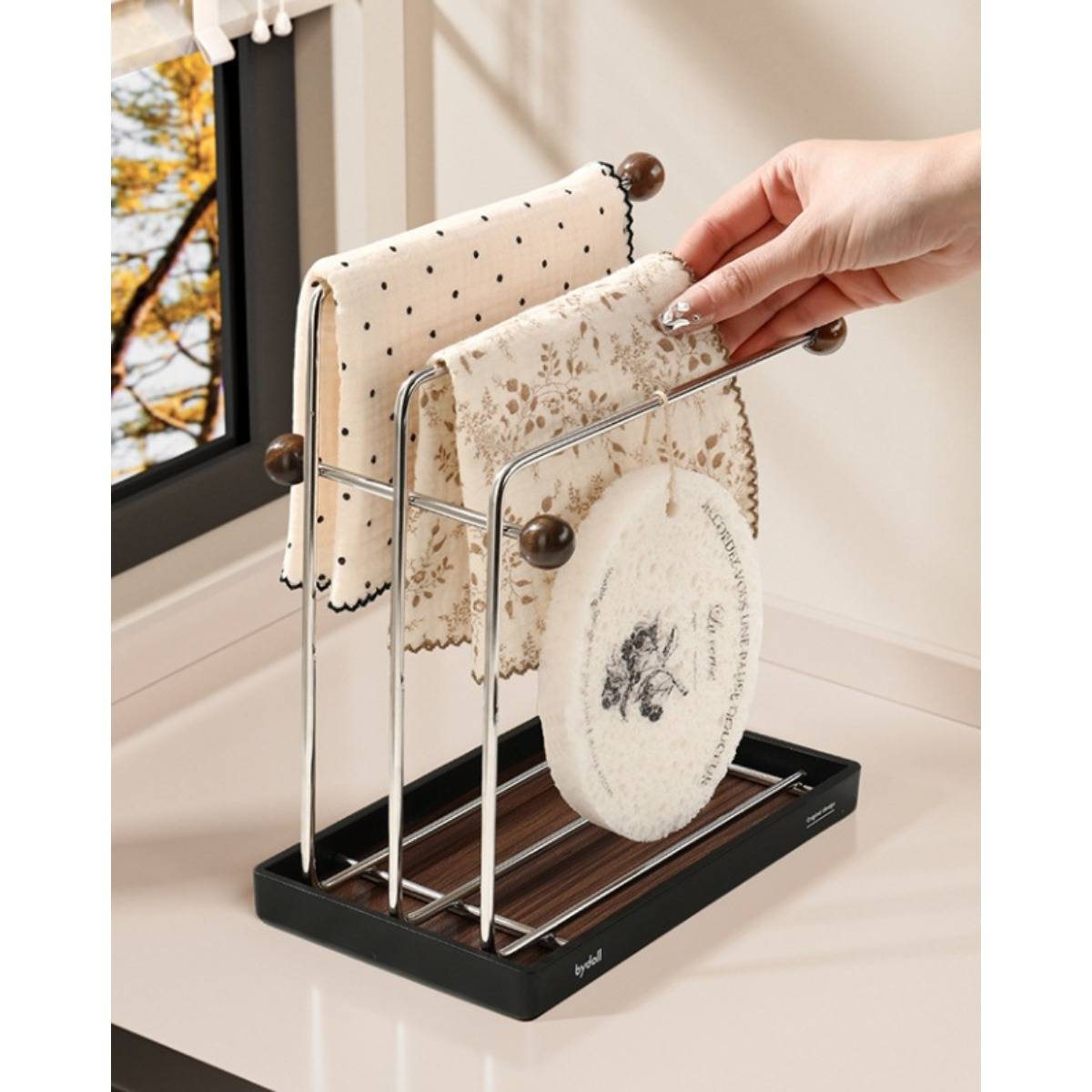 Eine Hand legt ein dekoratives Geschirrtuch auf einen Metallzähler-Handtuchhalter, der zwei Handtücher und ein rundes Tuchpad neben einem Fenster hält.