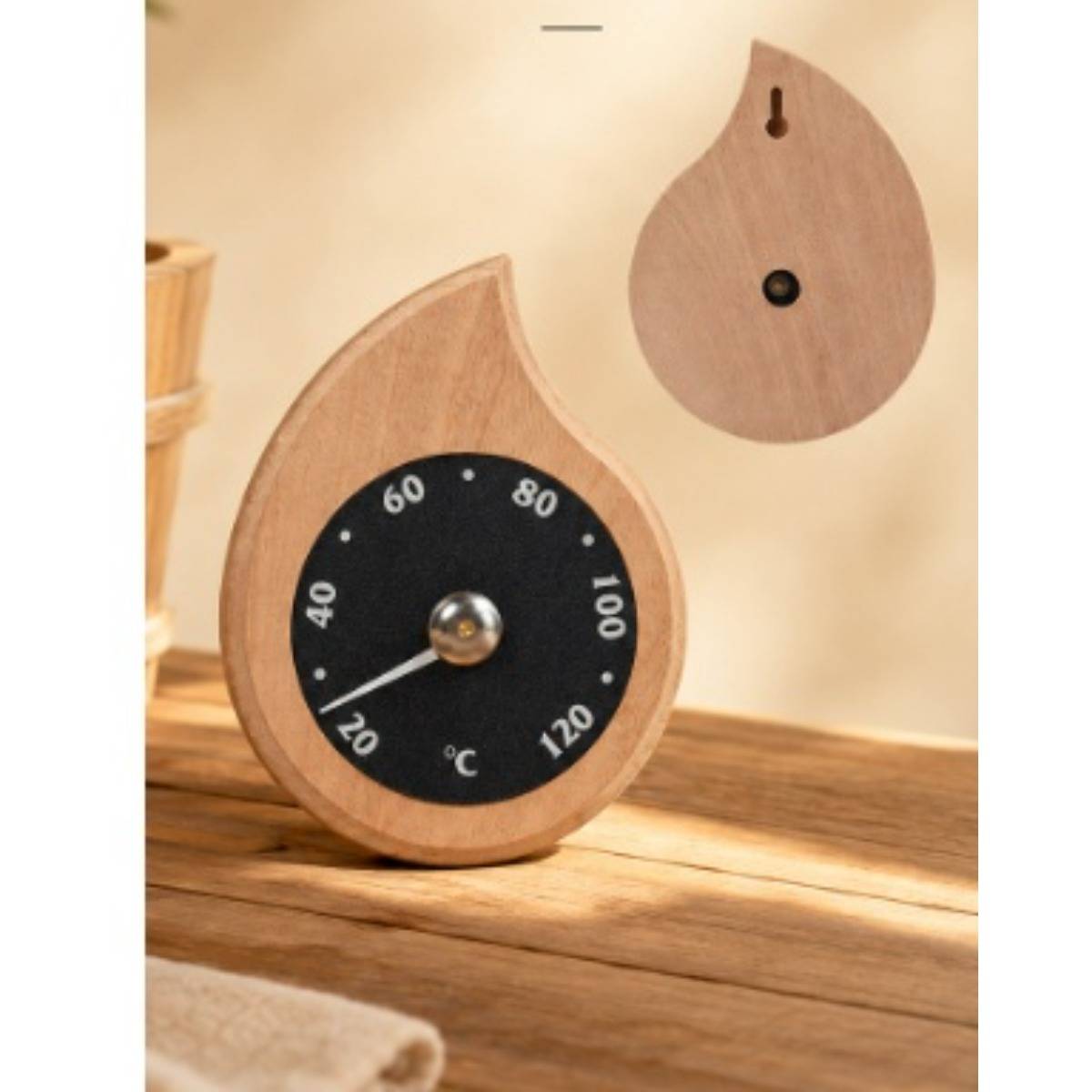 Holzförmiges Thermometer in Tränenform auf einer Holzoberfläche, das ein schwarzes Zifferblatt mit weißen Celsius-Markierungen von 20 bis 120 und eine Nadel in der Nähe von 20°C zeigt; eine passende Rückseite hängt oben darüber.