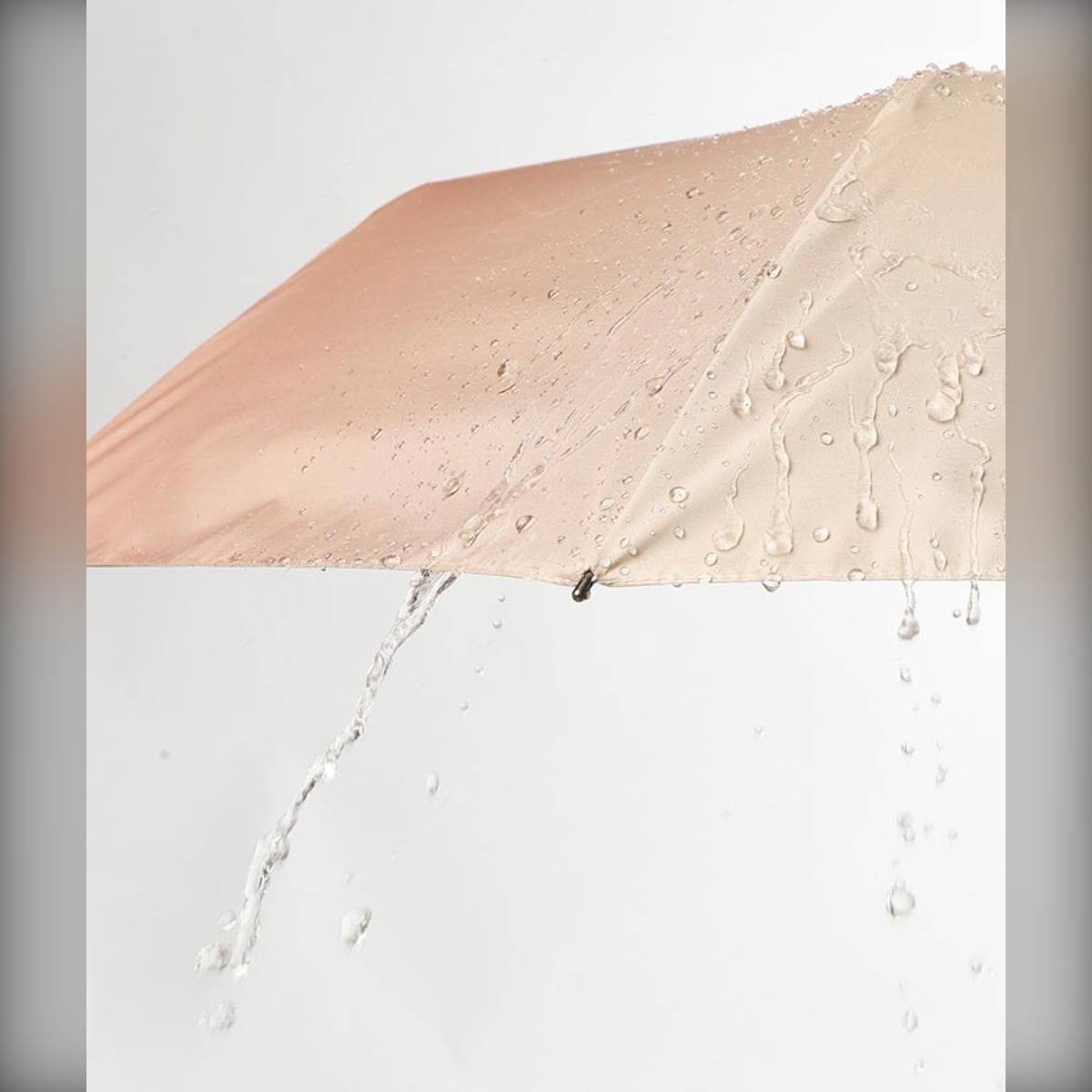 Nahaufnahme eines beigefarbenen Regenschirms, von dem Wasser abperlt, mit Regentropfen, die auf seiner Oberfläche perlen und herabfließen.