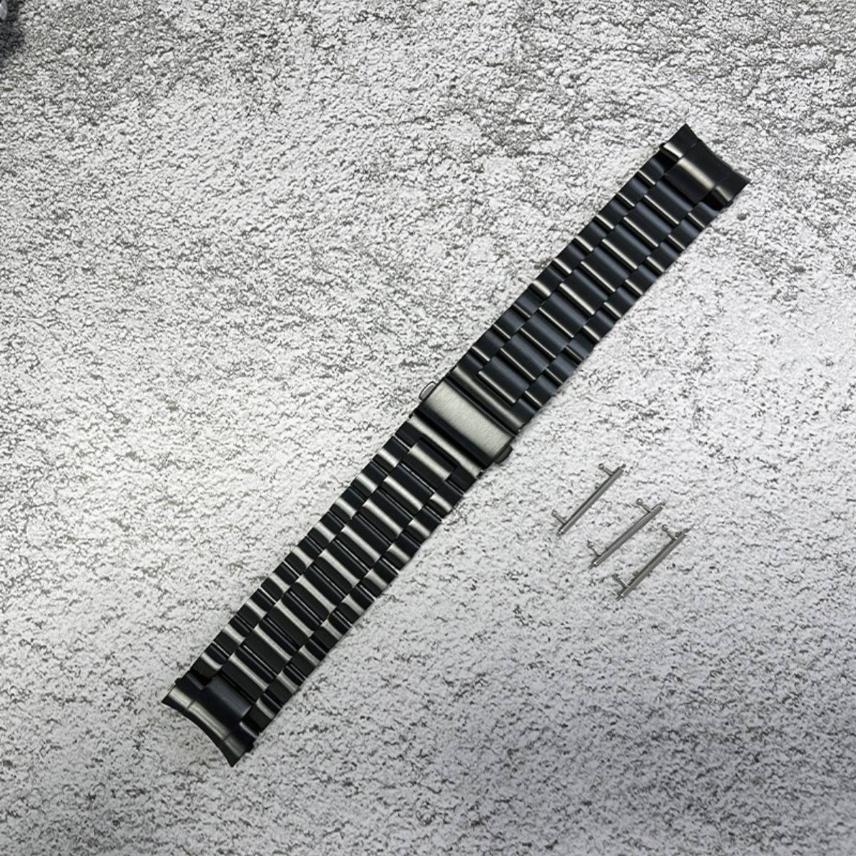 Schwarzes Metalluhrenarmband diagonal auf einer strukturierten grauen Oberfläche angeordnet, mit drei kleinen Einstellstiften daneben.