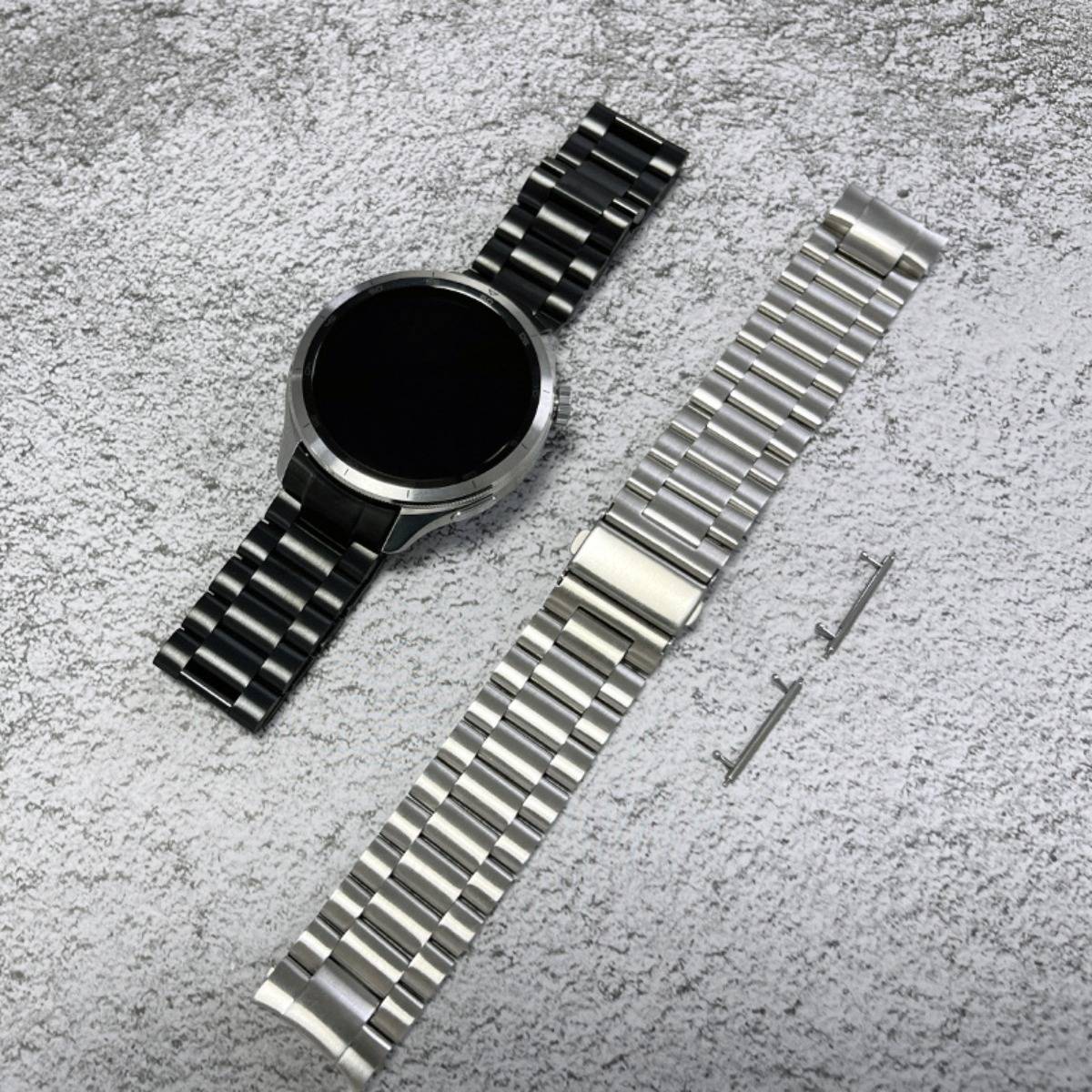 Rundige Smartwatch mit schwarzem Metallarmband, dargestellt neben einem separaten silbernen Austauscharmband und zwei Verbindungsstiften auf einer strukturierten Oberfläche.